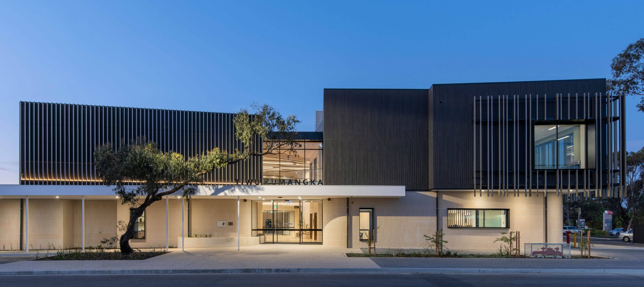 Tiwu Kumangka at dusk. The warm sandstone blockwork and dark timber-look upper level glows with architectural lighting. The building name is black above the entry canopy. A gum tree reaches accross the front entryway. Strong vertical fins wrap the upper level.